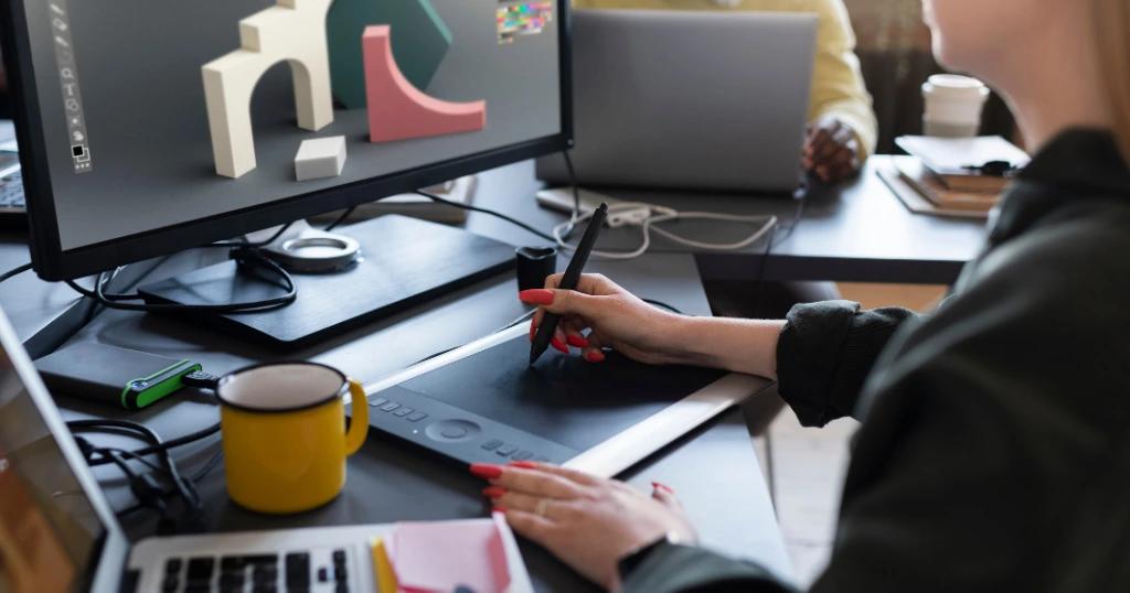 A graphic designer using a digital pen tablet to create 3D shapes on a computer monitor, surrounded by other team members at work.