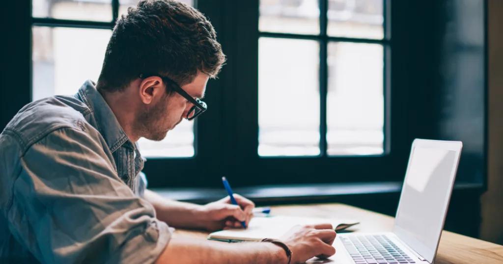 Content writer in glasses working on laptop, taking notes in modern office space. Writing agency professional focused on creating content.