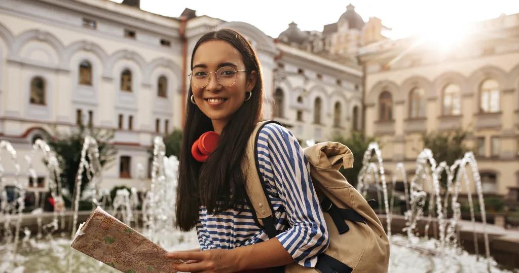 A young woman holding a map, smiling near a fountain, preparing for her UK Visa journey.