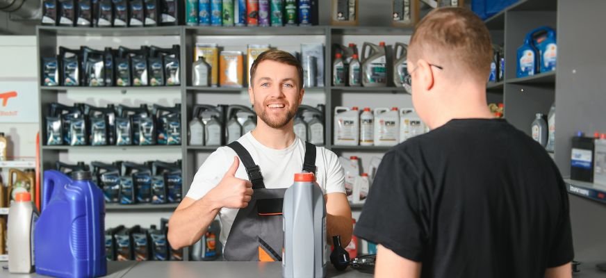 Smiling worker at a petroleum retail store offering oil products to a customer, with shelves of lubricants in the background.